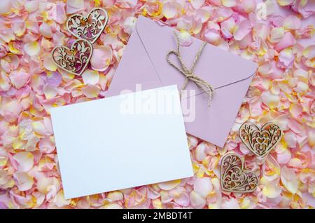 Gestalte einen rosafarbenen Umschlag mit unbeschrifteter Karte, dekorativen Holzherzen, Rosenblättern und Knospen auf pinkfarbenem Strickhintergrund. Hintergrund zum Valentinstag und Copy Spac Stockfoto