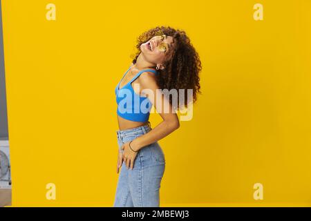 Eine Frau mit lockigem Afro-Haar in einem blauen T-Shirt auf gelbem Hintergrund tanzt fliegende Haare mit gelber Sonnenbrille, Handzeichen, schauen Sie in die Kamera Stockfoto