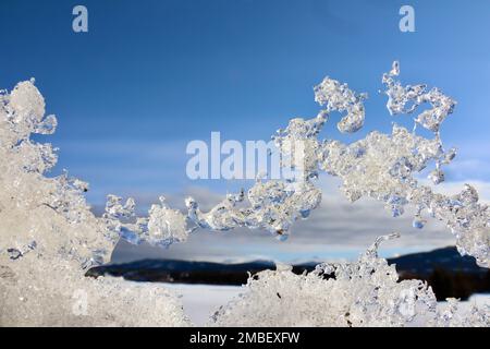 Nahaufnahme von tanzenden Eisfiguren, die Frost erleben und dann bei Sonnenschein auftauen, mit blauem Himmel, gefrorenem Fluss und Bergen im Hintergrund Stockfoto