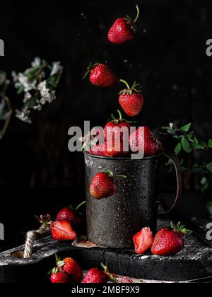 Nahaufnahme eines nassen Jahrgangs mit Erdbeeren auf einem Holzbrett, umgeben von herunterfallenden Erdbeeren, auf schwarzem Hintergrund Stockfoto