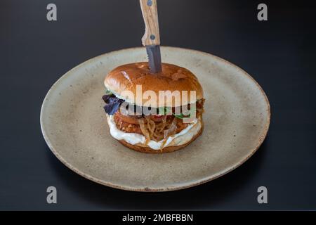 Hamburger-Cheeseburger mit Schweinehähnchen-Käse auf einem Teller und einem steckengebliebenen Messer. Stockfoto