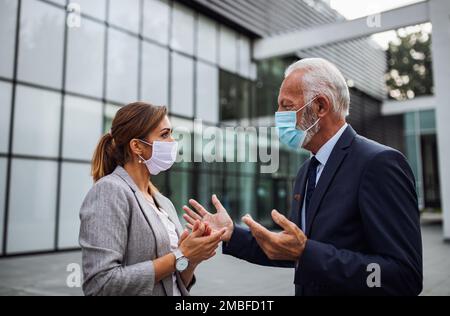 Geschäftsmann und Geschäftsfrau tragen Sicherheitsmasken und sprechen vor dem Büro. Soziale Distanzierung und Virenschutzkonzept Stockfoto
