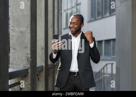 Erfolgreicher afroamerikanischer Boss außerhalb des Bürogebäudes, der telefonische Geschäftsmann im Anzug nutzt, um den Sieg zu feiern, erfolgreiche Erfolge, Mann, der Hand hoch hält, Triumph-Geste, Online-Benachrichtigung liest Stockfoto