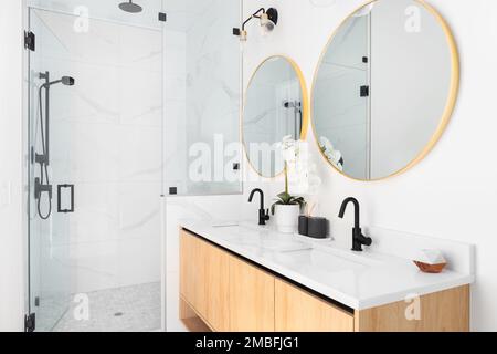 Ein wunderschönes Badezimmer mit einem schwimmenden Schrank aus Holz, runden goldenen Spiegeln, einer Marmorablagefläche und einem mit Marmor ausgekleideten Spiegel mit schwarzen Wasserhähnen. Stockfoto