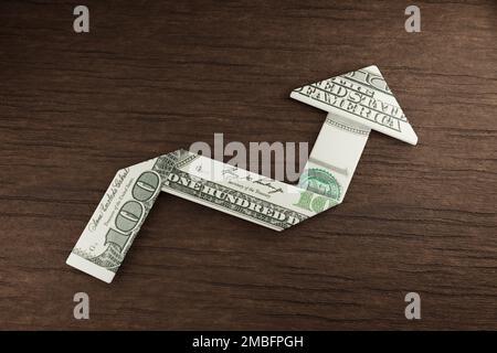 100-Dollar-Schein, gefaltet in einem Pfeil nach oben auf einem Holztisch. Veranschaulichung des Konzepts von Inflation, Gewinn, Ertrag und Investition Stockfoto