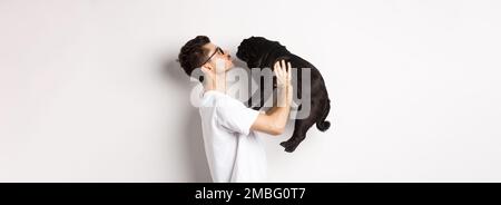 Profil von schönen jungen Mann küssen kleinen niedlichen Hund Gesicht. Hipster Kerl liebt seinen Mops, stehen über weißem Hintergrund Stockfoto
