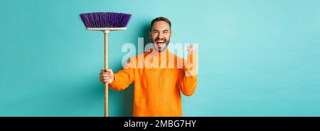 Bild von ermutigt und motiviert Mann immer bereit für die Reinigung, hält Besen und macht Faust Pumpe Geste, stehen über türkisfarbenem Hintergrund Stockfoto