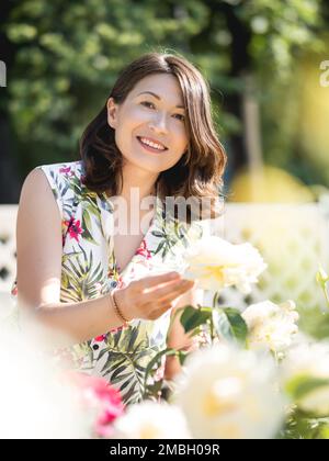 Eine weiße Frau bewundert blühende Rosen im öffentlichen Park. Sommerliche Stimmung. Tropische Pflanzen und Blumen blühen im Garten. Stockfoto