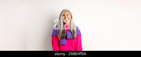 Lustige und glückliche asiatische ältere Frau mit Spaß, zeigt Zunge und winken frech, Blick links auf Logo, stehend über weißem Hintergrund Stockfoto