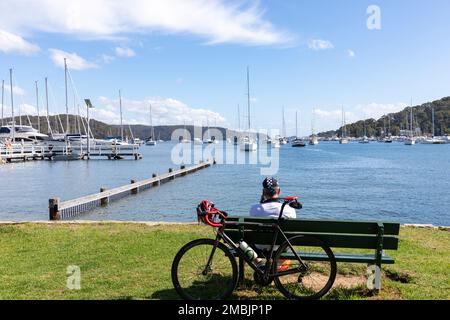 Männlicher Radfahrer macht eine Pause Bayview Sydney Australia auf einer Parkbank und genießt den Blick über Pittwater und den Jachthafen im Sommer 2023 Stockfoto