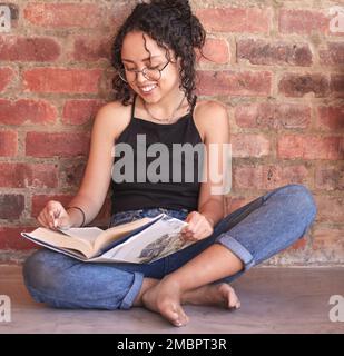 Je mehr ich lese, desto mehr lerne ich. Eine junge Frau, die ein Buch liest, während sie an einer Ziegelwand sitzt. Stockfoto