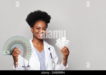 Erfolgreiche afroamerikanische Ärztin gp lächelt und hält Fans von Geld und Päckchen Pillen vor grauem Hintergrund Stockfoto