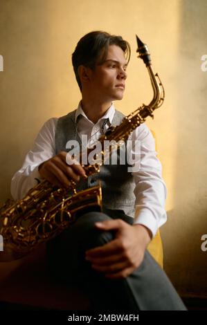 Junger Mann, der mit Saxophon auf einem Stuhl sitzt Stockfoto