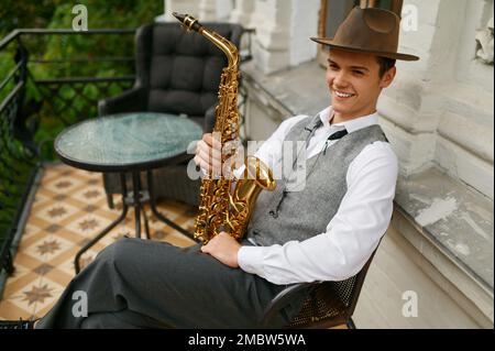 Lächelnder Musiker mit Saxophon, der auf einem Stuhl auf dem Balkon seiner Wohnung saß Stockfoto