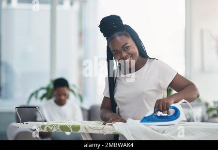 Der Mann kümmert sich um das Geschäft, ich kümmere mich um die Hausarbeit. Eine junge Frau bügelt Kleidung mit ihrem Mann auf dem Sofa im Hintergrund. Stockfoto