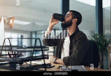 Den Kaffee zurückschlagen, um die Uhr zu schlagen. Ein junger Geschäftsmann, der in der späten Nacht in einem modernen Büro Kaffee trinkt und einen Computer benutzt. Stockfoto