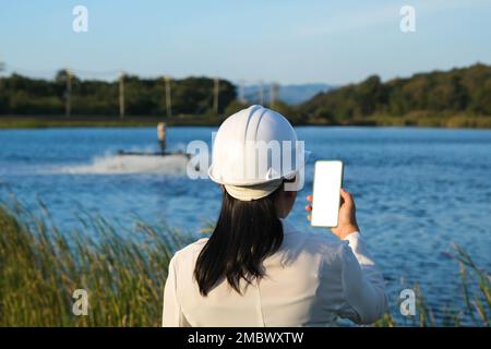 Ein Umweltingenieur, der einen weißen Helm trägt, verwendet ein Mobiltelefon, um einen Sauerstoffturbinenlüfter in einem Teich zu betreiben. Wasser- und Ökologiekonzept. Stockfoto