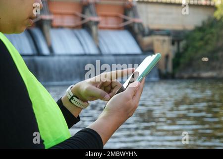 Ingenieurin in White hat, die mit Smartphone arbeitet und mit Wasserkraftwerk und Bewässerung auf den Damm blickt. Stockfoto