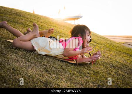 Glückliche Schwestern, die im Park spielen, rutschen vom Grashügel hinunter, sitzen auf einem Karton. Fröhliche Kinder spielen im Sommer draußen. Familienausgaben Stockfoto