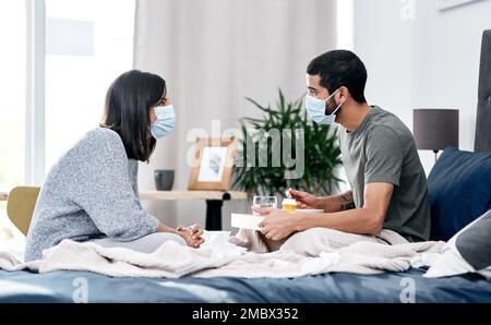 Manchmal meine Krankenschwester, immer meine Frau. Ein maskiertes junges Paar, das im Bett frühstückt, während es sich von einer Krankheit zu Hause erholt hat. Stockfoto