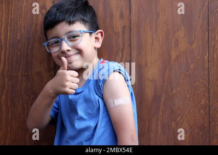 Der kleine 6-jährige Latino-Junge mit Brille und blauem Hemd zeigt seinen Arm mit einem Verband, weil er gerade gegen Covid-19 geimpft wurde Stockfoto