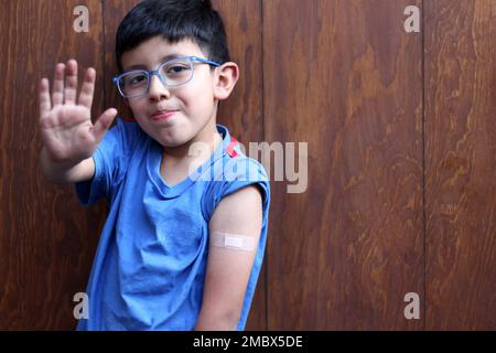 Der kleine 6-jährige Latino-Junge mit Brille und blauem Hemd zeigt seinen Arm mit einem Verband, weil er gerade gegen Covid-19 geimpft wurde Stockfoto