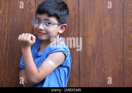 Der kleine 6-jährige Latino-Junge mit Brille und blauem Hemd zeigt seinen Arm mit einem Verband, weil er gerade gegen Covid-19 geimpft wurde Stockfoto