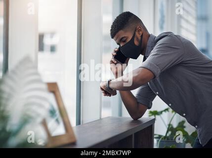 Wann kann ich mit einem Update rechnen? Ein junger Geschäftsmann, der eine Gesichtsmaske trägt und die Uhrzeit überprüft, während er in einem Büro mit einem Handy spricht. Stockfoto