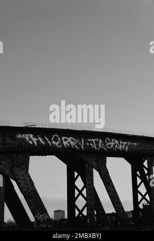 Ein vertikales Bild eines spritzgestrichenen Zitatzeichens auf einer hohen Metallbrücke in Graustufen Stockfoto