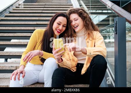 Zwei Frauen lächeln und haben Spaß, während sie ihr Handy benutzen und draußen auf der Treppe sitzen. Stockfoto