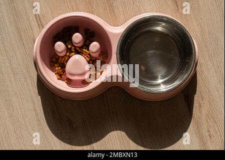 Eine Doppelschüssel für langsames Füttern und eine Schüssel Wasser für den Hund. Draufsicht auf einen rosafarbenen Teller mit trockenen Speisen auf einem Holzboden. Stockfoto