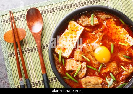 Das Sundubu-jjigae-Gericht wird mit frisch gerocktem, weichem Tofu, Kimchi, Zwiebeln, Schweinebauch und Gochugaru-Nahaufnahme auf der Schüssel auf dem Tisch zubereitet. Horizontal oben V Stockfoto