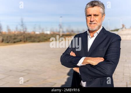 Seriöser, bärtiger Geschäftsmann in formellem Anzug, der auf der Straße steht, mit gekreuzten Armen und lächelt, während er in die Kamera schaut Stockfoto