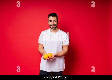 Porträt eines jungen bärtigen Mannes, der Zitronen in beiden Händen auf einem isolierten roten Hintergrund hält Stockfoto