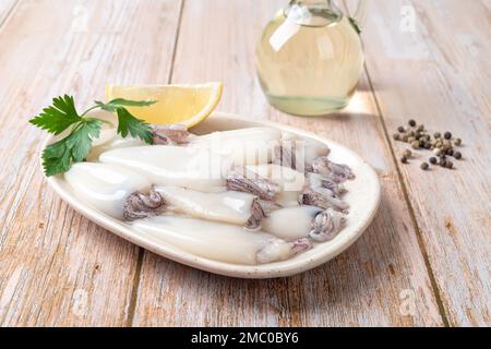 Frische Tintenfischröhren auf einem Teller über einem Holztisch. Kleines Kalamarfilet, Zitrone und Olivenöl zum Kochen zubereitet. Roher Tintenfisch für kalorienarmes Rezept. Stockfoto