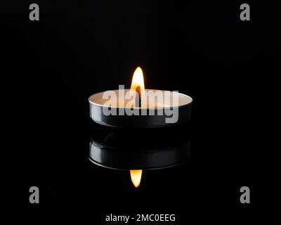 Brennende Teelichterkerze in der Dunkelheit. Die Kerze leuchtet auf einer schwarzen glänzenden Oberfläche. Konzentrieren Sie sich auf ein einzelnes Kerzenlicht vor schwarzem Hintergrund. Stockfoto