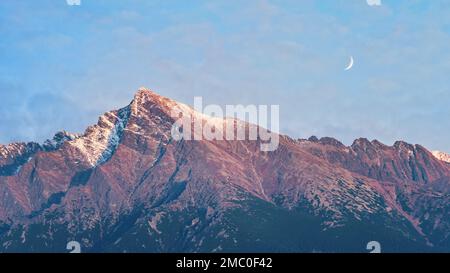 Der Halbmond erhebt sich über dem Gipfel von Krivan - slowakisches Symbol - Waldbaumsilhouetten im Vordergrund, Abendszene Stockfoto