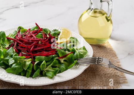 Lammsalat mit roher Beete. Grüne Maissalatblätter, geriebener roher Rüben und Gabel auf einem weißen Teller über der Marmoroberfläche. Kalorienarme Lebensmittel. Stockfoto