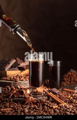 Der Kaffeelikör wird aus einer Flasche in ein Glas gegossen. Kaffeebohnen, Zimt, Anis und Stücke bitterer Schokolade sind auf dem Tisch verstreut. Stockfoto