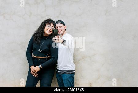 Ein junges Paar macht ein Selfie an einer Wand mit Kopierraum, junge Freunde macht ein Selfie, lehnt sich an eine Wand, ein junges glückliches Paar macht ein Selfie Stockfoto