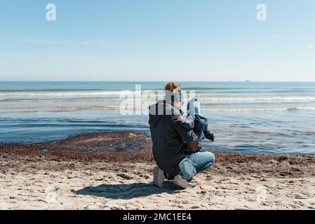 Rückansicht der Mutter, die ein Kleinkind am Strand hält, gegen das blaue Meer und den klaren blauen Himmel Stockfoto