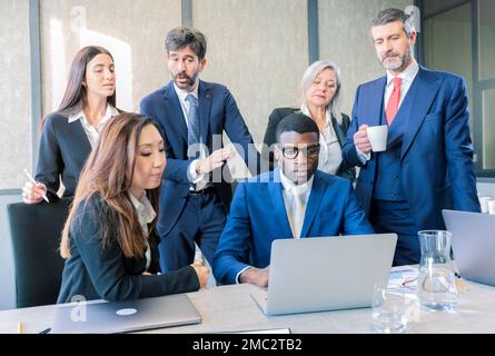 Eine Gruppe von Kollegen in formeller Kleidung, die einen Tisch mit Laptop-Computer, Kaffee zum Mitnehmen und Papierblätter und -Ordner tragen, die an einem offenen Ort von arbeiten Stockfoto