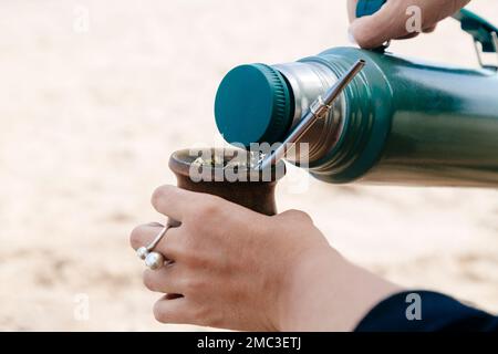 Nahaufnahme von Frauenhänden, die heißes Wasser servieren, einer Thermoskanne in einem Partner mit Yerba. Trinkender Kumpel am Strand. Stockfoto