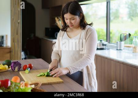 Schwangere Asiatin schneidet Gurke für frischen grünen Salat, weibliche bereitet leckeres Bio-Abendessen zu Hause zu, gesunde Ernährung für zukünftige Mutter Stockfoto