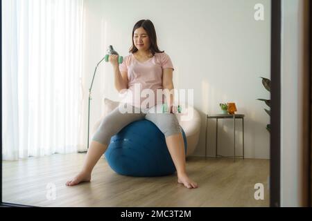 Schöne asiatische schwangere Frau, die zu Hause mit Kurzhanteln trainiert, gesundes Konzept Stockfoto