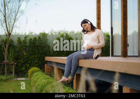 Porträt einer schwangeren Happy Asian Woman, die zu Hause auf der Terrasse sitzt und über ein Smartphone Musik über Kopfhörer hört. Stockfoto