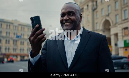 Lächelnder afroamerikanischer männlicher Mann mittleren Alters Arbeitgeber Unternehmer Unternehmer in der Stadtstraße draußen sprechen Handy Kamera Talk Stockfoto