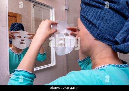 Porträt einer Frau, die nach der Dusche in ihrem Badezimmer eine Gesichtsmaske aus Baumwolle aufträgt. Pflege der Hautschönheit. Teint-Behandlungskonzept. Stockfoto
