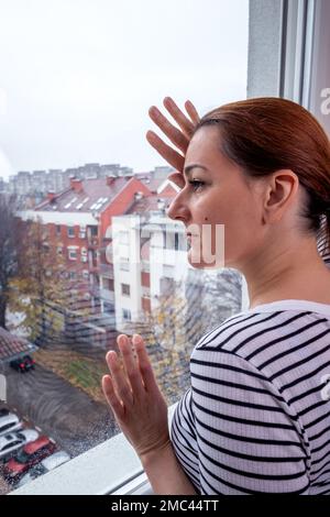 Eine Frau, die durchs Fenster schaut. Junge Frau zu Hause, mit einem traurigen Blick und ihrem Kopf und ihren Händen auf dem Fensterglas. Depression. Psychische Gesundheit. Stockfoto