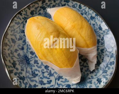 Zwei goldgelbe Fleischsorten von Durian, eingewickelt mit weißem Papier auf dem blauen, antiken Teller, König der Früchte, natürliche Form und Form, dunkle Hintergrundfarbe Stockfoto